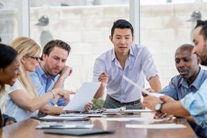 Man standing at end of conference table talking to a group of professionals looking at papers on the table.